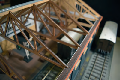 

There is a canopy to the road side and this fits on five brackets. The
roof fits onto this: ‘teeth’ and a build-up of strip-wood supplied form the
final saw-tooth valancing.
There are six substantial roof A-frame trusses which sit on the side
wall plates with a ridge board full length. The roof sits on the trusses and
is fixed on one side only. The ridge is formed from plastic half round fixed
to one edge only and used as a locating piece. With hindsight a thinner
right-angled extrusion would be better and I will replace it shortly. All
my roofs are fixed in a similar way. An alternative would be to build the
roof as a totally detachable unit by not gluing the trusses onto the wall
plates, but this could make any lighting difficult to install other than on
full width dummy beams. All roofs are usefully scribed to assist with the
alignment of the paper tiles. Again my preference is for double-sided
tape. The chimneys are themselves a work of art and fit well and locate
onto the trusses.

{{:gazettearchive:gazettevol21:tetbury_shed_10.png?400|